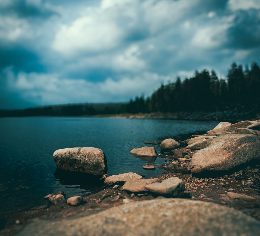 Steine am Ufer des Oderteichs, dahinter See und Wald unter dunklen Wolken